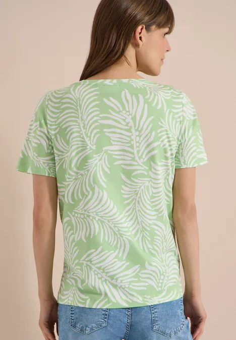 Rugaanzicht van een vrouw met een lichtgroen t-shirt met wit bladerenpatroon en een blauwe spijkerbroek.