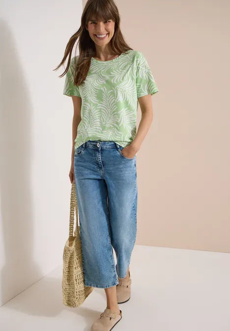 Vrouw in groen-wit patroon-T-shirt, wijd jeans, strozak en sandalen.