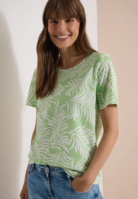Vrouw met groen T-shirt met wit bladerenpatroon en spijkerbroek, handen in de zakken.