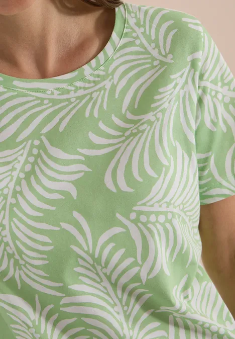 Vrouw in lichtgroen t-shirt met wit palmbladpatroon, alleen bovenlichaam zichtbaar.