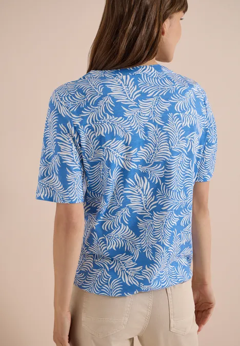 Frau von hinten in blauem T-Shirt mit weißem Blattmuster und beiger Hose.
