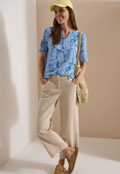 Frau in blau gemustertem Kurzarmshirt, weiter beiger Hose, mit Basttasche und Loafern vor schlichter Kulisse.