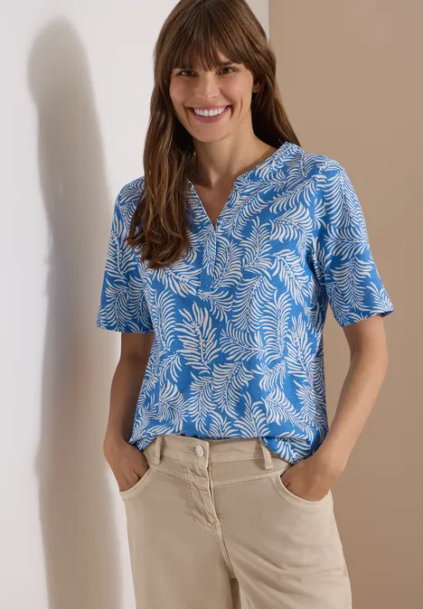 Frau in blau-weiß gemustertem Kurzarmshirt und heller Hose, Hände in den Taschen, vor beigem Hintergrund.