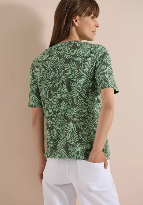 Rückansicht einer Person mit langen Haaren trägt ein grünes T-Shirt mit Blattmuster und weiße Hose.