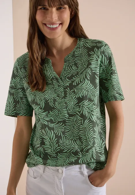 Frau mit grünem Blattmuster-Shirt und weißer Hose, Hände in den Hosentaschen.