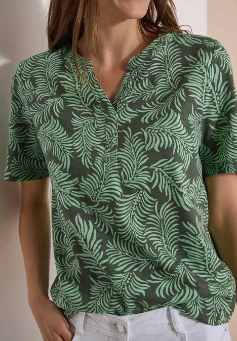 Frau in grünem V-Shirt mit Blattmuster und weißen Hosen.