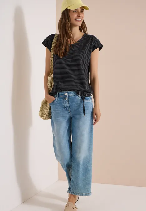 Eine Frau in schwarzem T-Shirt, weiten Jeans, beiger Strotasche und Cap vor hellem Hintergrund.