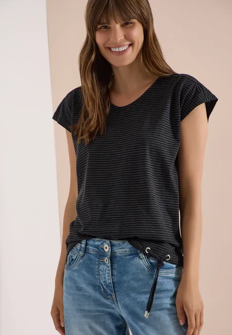 Eine Frau trägt ein dunkel gestreiftes T-Shirt mit Flügelärmeln und eine blaue Jeans mit Gürtel.