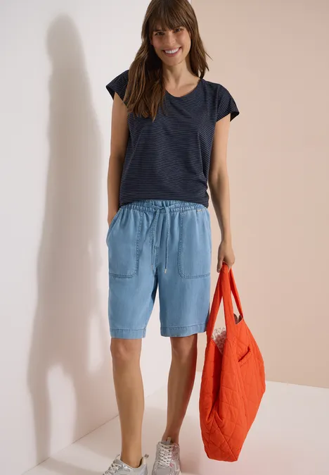 Person trägt dunkles T-Shirt, hellblaue Bermudashorts und hält eine orangefarbene Stofftasche.
