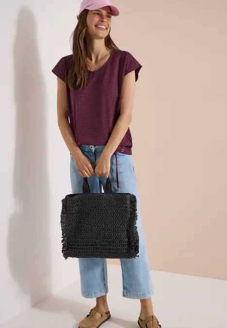 Frau in lila T-Shirt und Jeans steht mit schwarzer Tasche in einer hellen Umgebung.