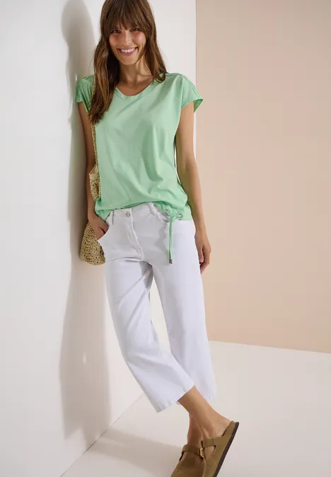 Femme en t-shirt vert et pantalon blanc, avec sac en paille et sandales, appuyée contre un mur clair.