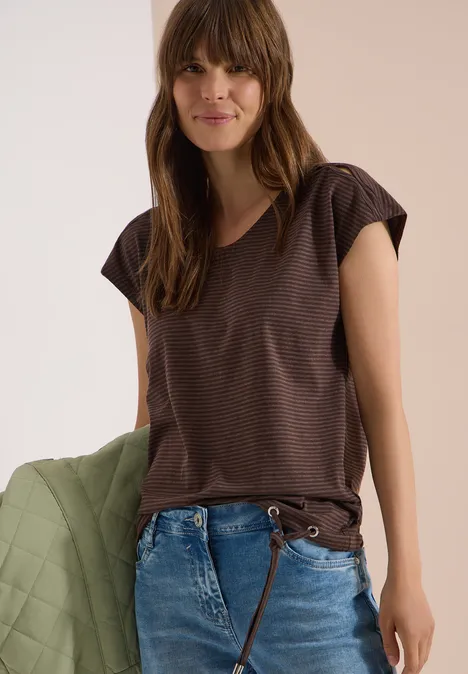 Frau trägt braunes, gestreiftes Kurzarmshirt mit V-Ausschnitt, blaue Jeans und hält eine gesteppte grüne Jacke.