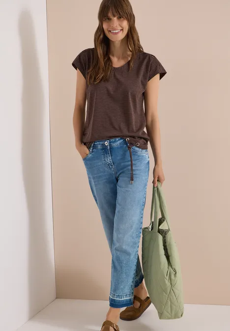 Frau vor hellem Hintergrund, trägt braunes T-Shirt, blaue Jeans und hält eine grüne Steppjacke.