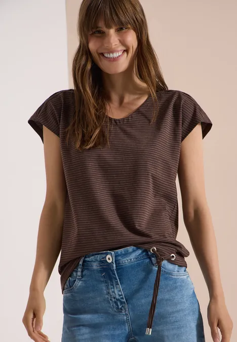 Frau in braunem Kurzarmshirt und blauen Jeans vor hellem Hintergrund.