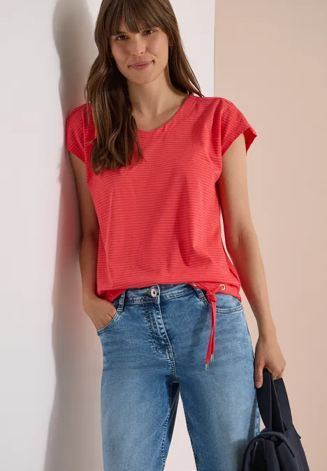 Femme en t-shirt rouge noué, jean bleu, tenant un sac foncé, debout devant un fond clair.