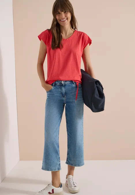 Femme en t-shirt rouge, jean évasé court et baskets blanches, portant une veste bleu marine.