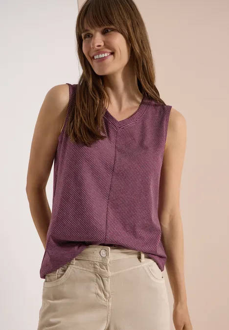 Top met V-hals en strepen sweet grape red Een vrouw draagt een paars mouwloos shirt en beige broek voor een neutrale achtergrond.