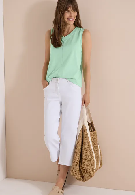 Top met V-hals en strepen juicy lime Vrouw in groen hemd, witte cropped jeans en strooien tas voor een beige muur.