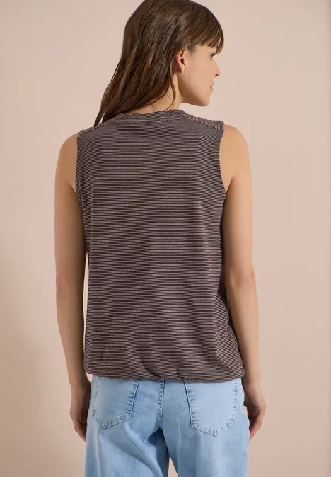 Junge Frau von hinten, trägt ärmelloses dunkelgraues Top und hellblaue Jeans, stehend vor beigem Hintergrund.