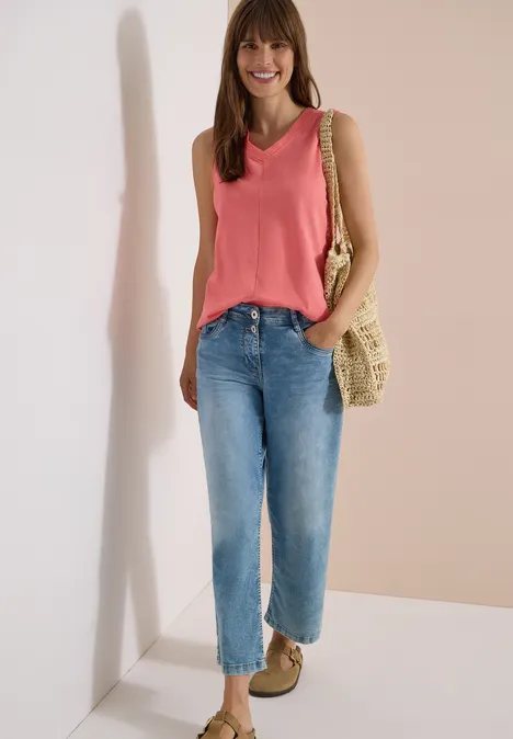 Top met V-hals en strepen coral Vrouw in roze mouwloos topje, blauwe jeans, strooien tas en beige schoenen, staand voor een neutrale achtergrond.