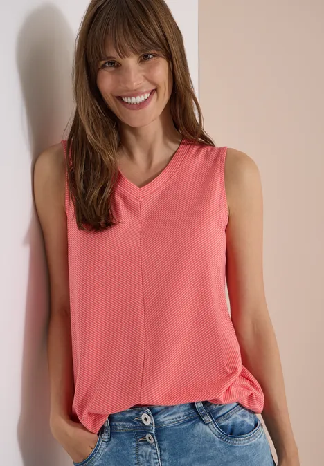 Top met V-hals en strepen coral Vrouw in een koraalkleurige V-hals top en jeans leunt ontspannen tegen een lichte muur.