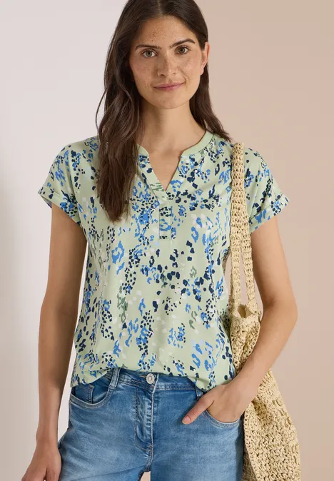 Frau trägt ein hellgrünes Kurzarmshirt mit blauem Blumenmuster, Knopfleiste, Jeans und Strohtasche.