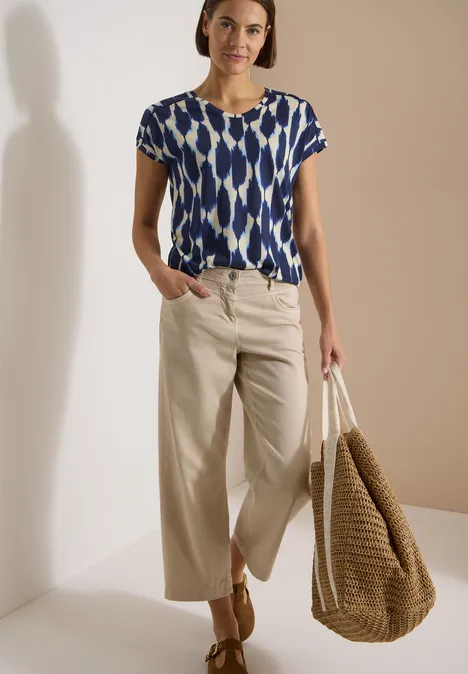 T-Shirt mit V-Neck und Spitzendetail urban dark blue Frau trägt blau-weiß gemustertes Shirt, beige Culotte-Hose, Sandalen und großen geflochtenen Strandkorb-Tasche
