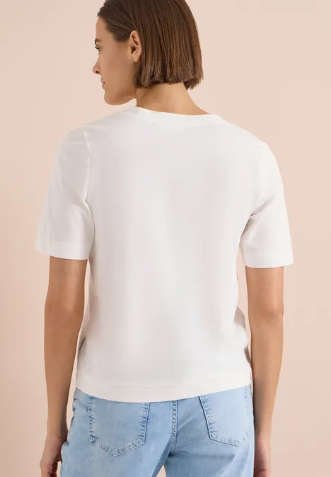 Shirt met V-hals en tekstdetail vanilla white Vrouw gezien van achteren met wit T-shirt en lichte spijkerbroek voor een beige muur.