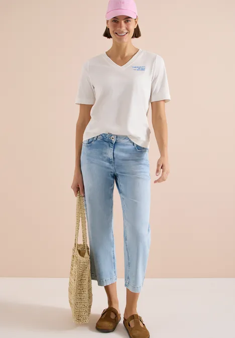 Shirt met V-hals en tekstdetail vanilla white Vrouw in wit V-hals T-shirt, lichtblauwe cropped jeans, bruine loafers en gevlochten strootas voor beige achtergrond.