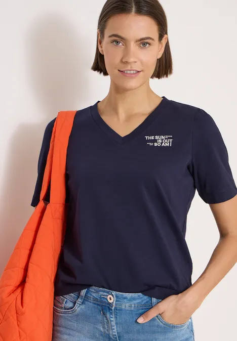 Shirt met V-hals en tekstdetail urban dark blue Vrouw draagt marineblauw V-hals t-shirt met wit logo, spijkerbroek en oranje schoudertas.