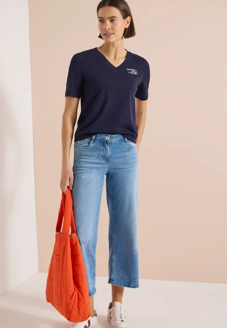 Shirt met V-hals en tekstdetail urban dark blue Vrouw met donkerblauw shirt, wijd lichtblauwe jeans, witte sneakers en een oranje tas voor een beige achtergrond