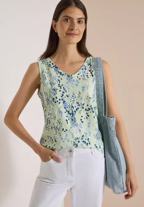 Junge Frau trägt ärmelloses, pastellgrünes Blumen-Top, weiße Hose und Tasche, steht vor heller Wand.