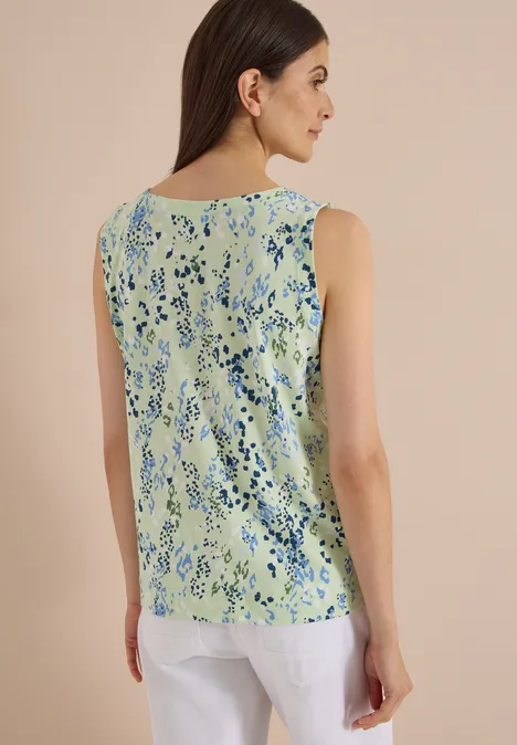 Frau von hinten in ärmellosem, hellgrünem Top mit blauem Blumenmuster und weißen Hosen.