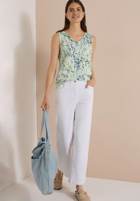Frau trägt geblümtes Top, weiße weite Hose und große blaue Tasche, steht vor neutralem Hintergrund.