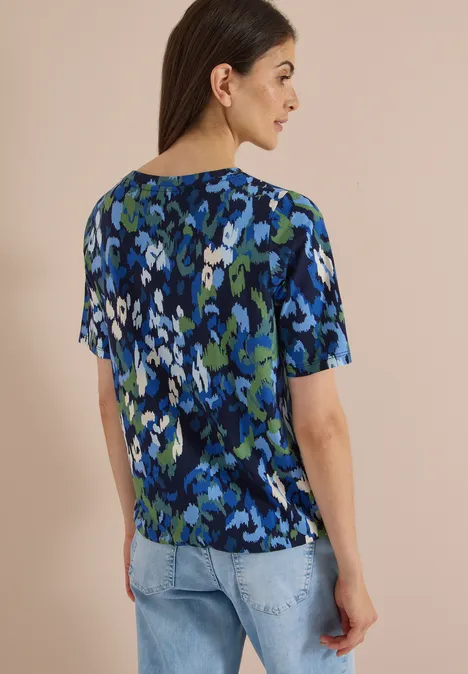Eine Frau steht seitlich, trägt eine kurzärmlige Bluse mit blau-grünem, abstraktem Blumenmuster und helle Jeans.