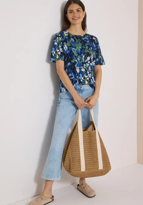 Frau trägt geblümtes blau-grünes Top, helle Schlagjeans, Strohtasche und beigen Clogs.