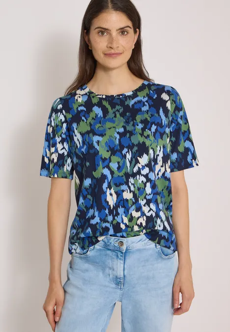 Frau in blau-grün gemusterter Bluse und hellen Jeans vor weißem Hintergrund.