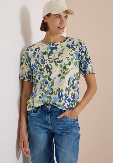 Vrouw draagt een bloes met bloemenprint, jeans en een crèmekleurige pet voor een lichte achtergrond.