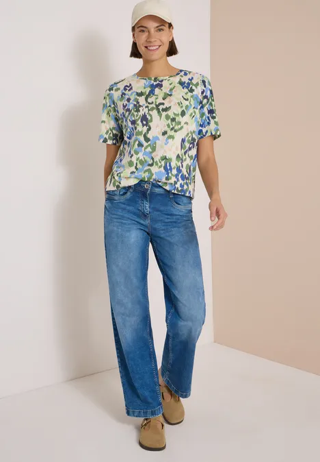 Vrouw draagt een blouse met bloemenmotief, wijde jeans en beige schoenen, glimlachend voor een neutrale achtergrond.