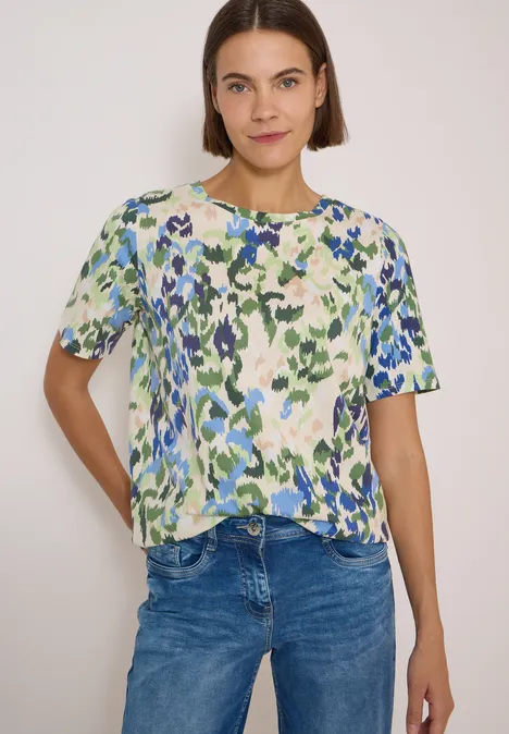 Vrouw in kleurrijk T-shirt met groen-blauwe abstracte bloemenprint en spijkerbroek.