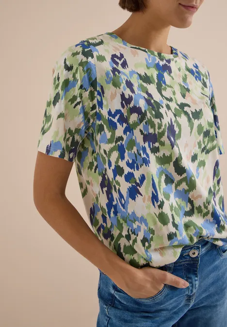 Vrouw draagt een shirt met korte mouwen en abstract bloemenpatroon in blauw en groen, gecombineerd met blauwe jeans.