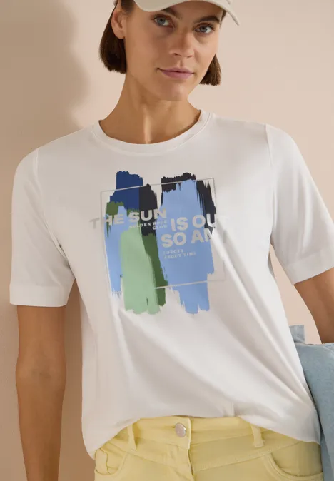 T-shirt blanc avec des traits bleu-vert et l'inscription 'THE SUN IS OUT SO AM I', associé à un pantalon jaune.
