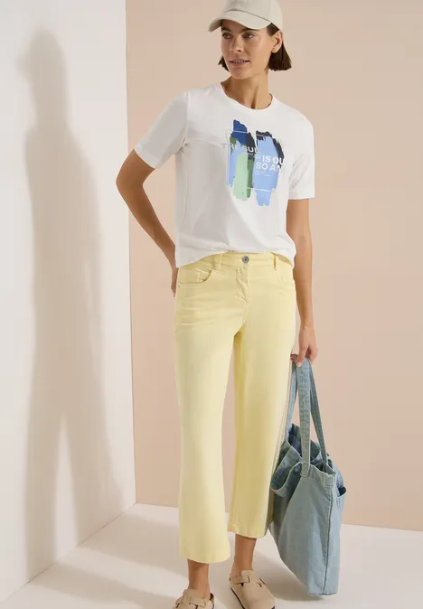 Femme portant un t-shirt blanc à motif abstrait, pantalon jaune et sac en jean bleu sur fond clair.