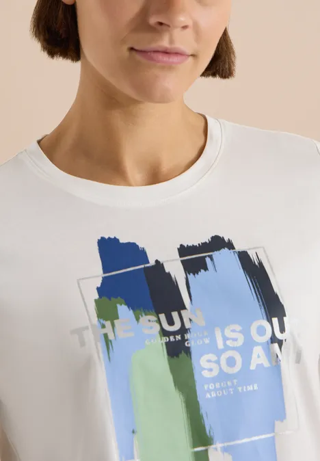 Femme au carré porte un t-shirt blanc orné de coups de pinceau abstraits bleu et vert et d’un texte imprimé.