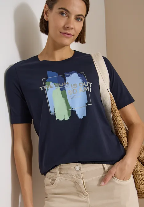 T-Shirt mit Rundhals und Frontprint urban dark blue Person trägt dunkelblaues T-Shirt mit pastellfarbenem Pinselstrich und Schriftzug, dazu beige Hose und Strohtasche.
