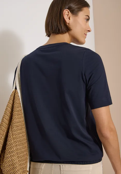 T-Shirt mit Rundhals und Frontprint urban dark blue Frau mit dunkelblauem T-Shirt, beige Hose trägt Strohtasche, Rückenansicht.