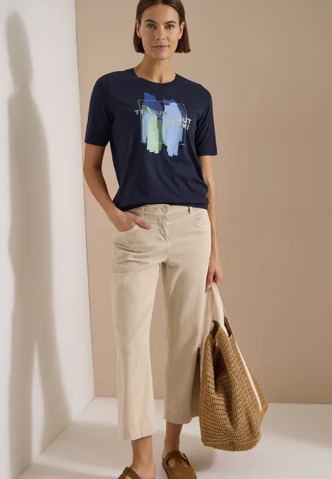 T-Shirt mit Rundhals und Frontprint urban dark blue Frau trägt blaues T-Shirt mit abstraktem Print, helle Culotte und Korbtasche vor neutralem Hintergrund.