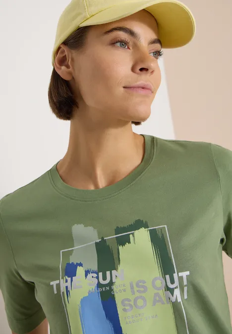 T-Shirt mit Rundhals und Frontprint deep matcha green Person mit gelber Cap und grünem T-Shirt mit Pinselstrich-Design und Text