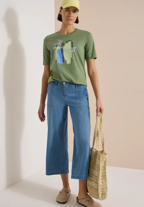 T-Shirt mit Rundhals und Frontprint deep matcha green Person trägt grünes T-shirt mit Print, weite blaue Culottes, Basttasche und beige Loafers.