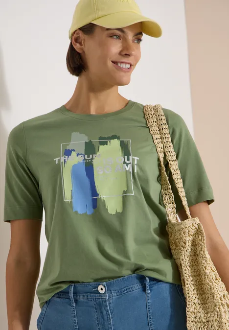 T-Shirt mit Rundhals und Frontprint deep matcha green Frau in gelbem Cap trägt grünes T-Shirt mit abstraktem Print, blaue Jeans und geflochtene Strohtasche.